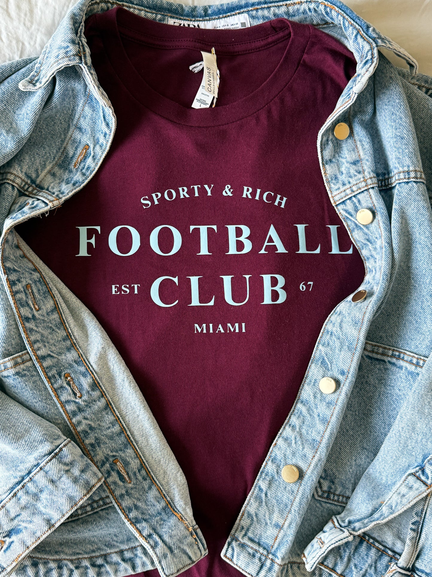 Maroon t-shirt with 'Sporty & Rich Football Club' text, worn over a denim jacket.