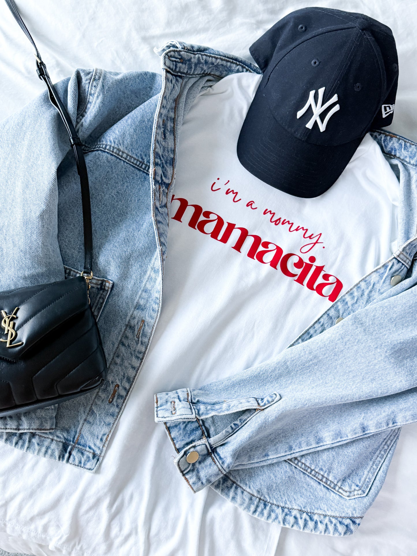 White t-shirt with red text, denim jacket, black cap, and black handbag on a light background