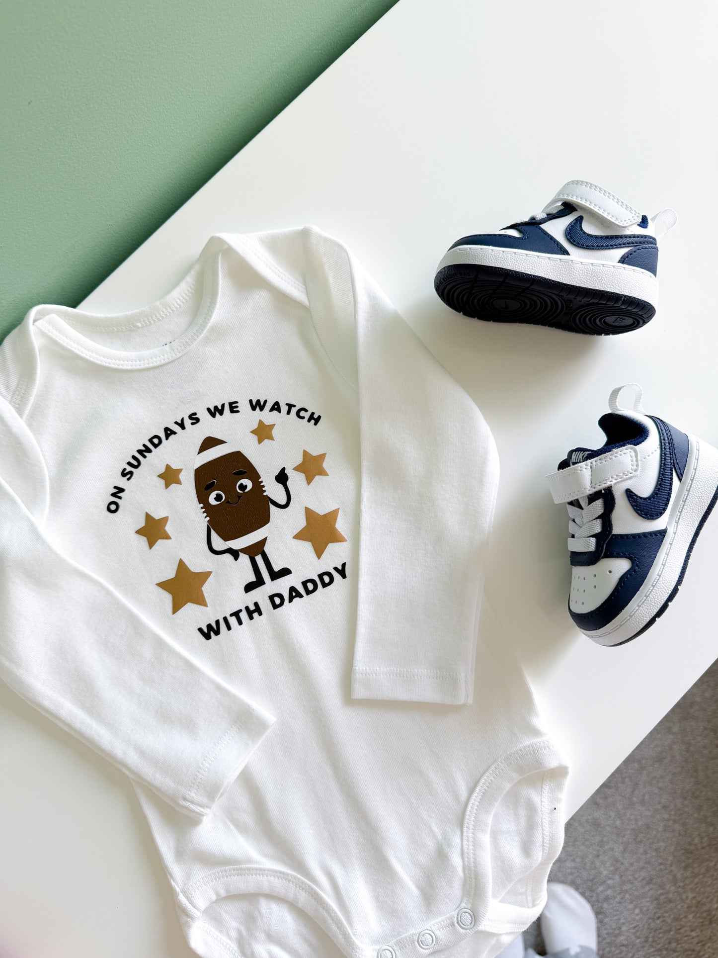 White baby onesie with a printed design and baby shoes on a light surface.