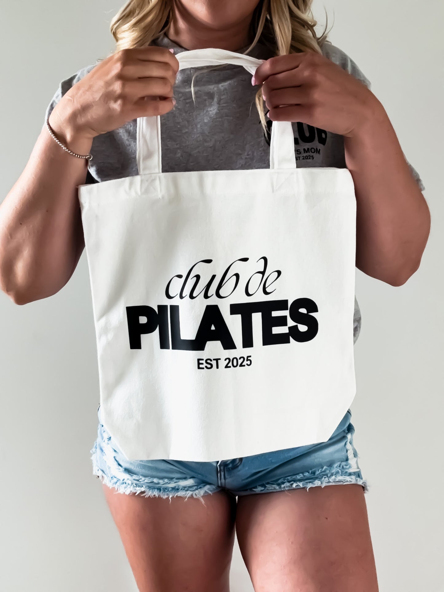 Person holding a tote bag with 'club de PILATES EST 2025' text on a plain background