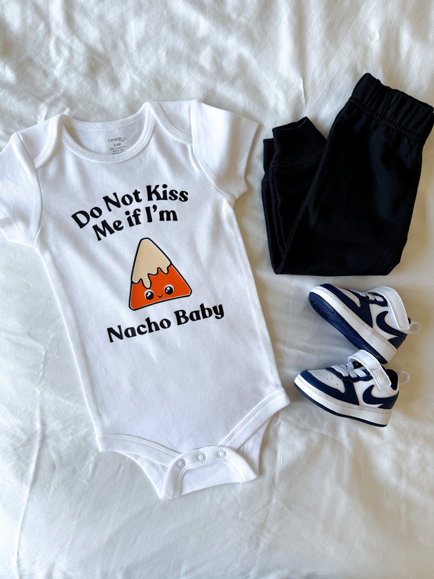 Baby onesie with 'Do Not Kiss Me if I'm Nacho Baby' text and nacho graphic, paired with black pants and blue sneakers on a white background.