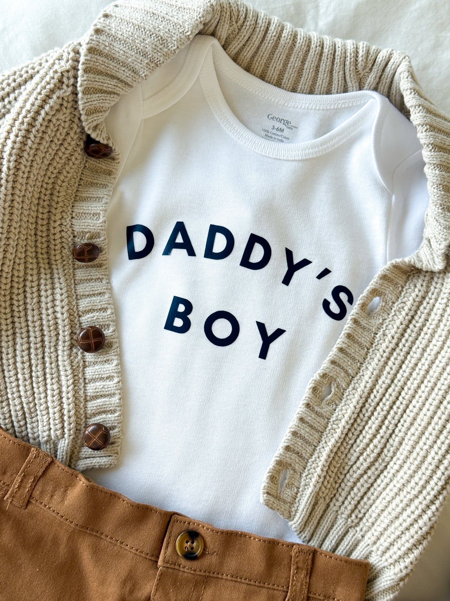 White baby onesie with 'DADDY'S BOY' text, beige knit cardigan, and brown pants on a light background