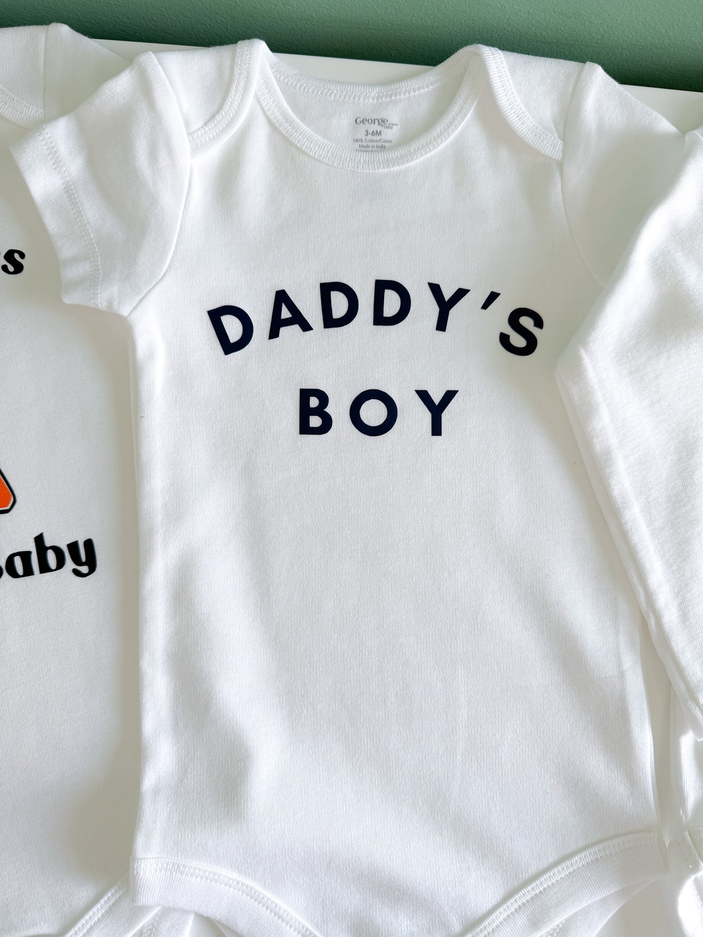 White baby onesie with 'DADDY'S BOY' text on a white background