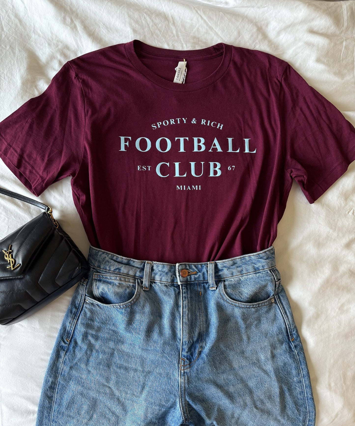 Maroon t-shirt with 'Football Club' text and blue jeans on a white surface