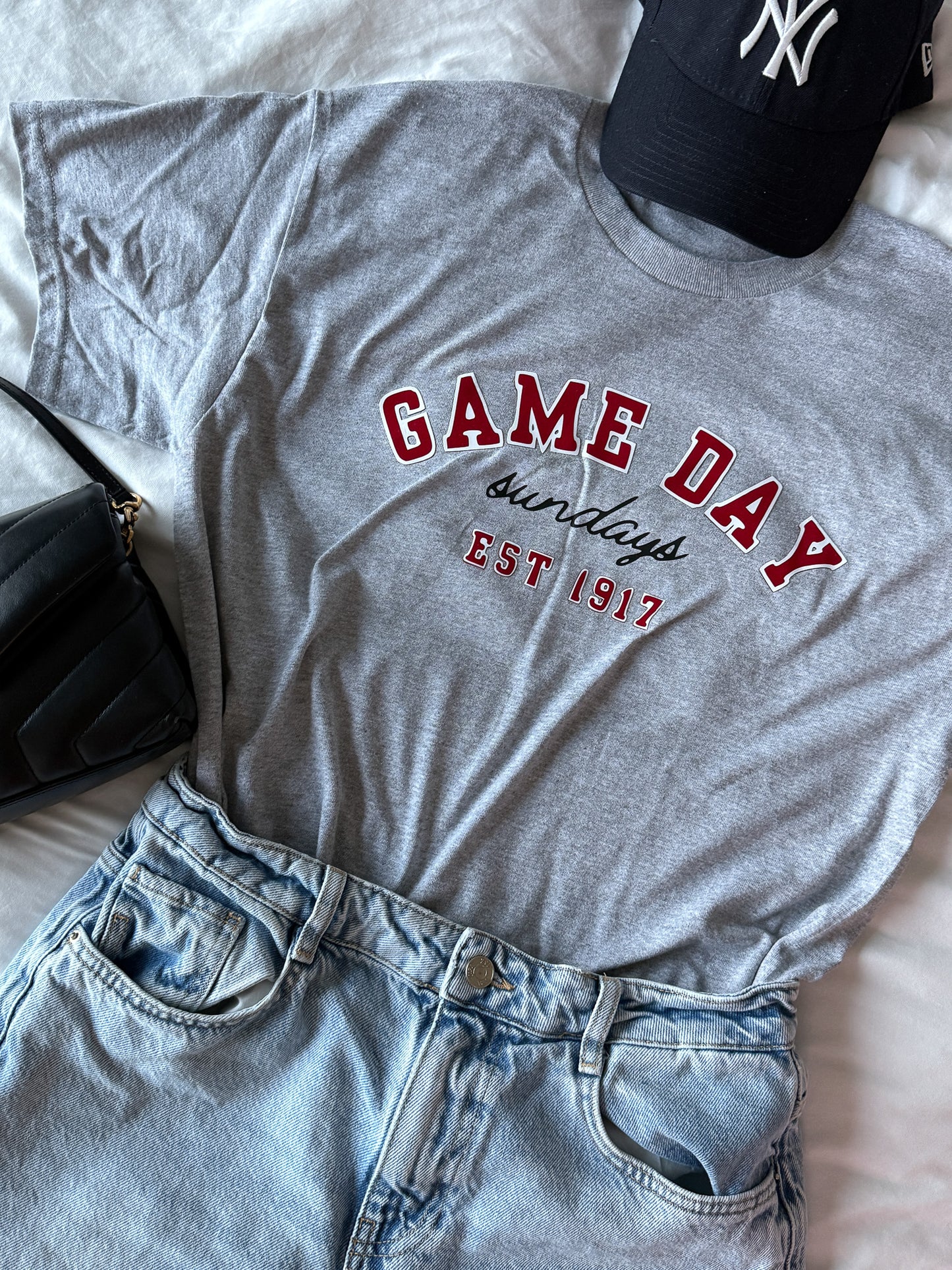 Gray t-shirt with 'Game Day Sundays EST 1917' text, paired with denim shorts and a black cap on a light background.