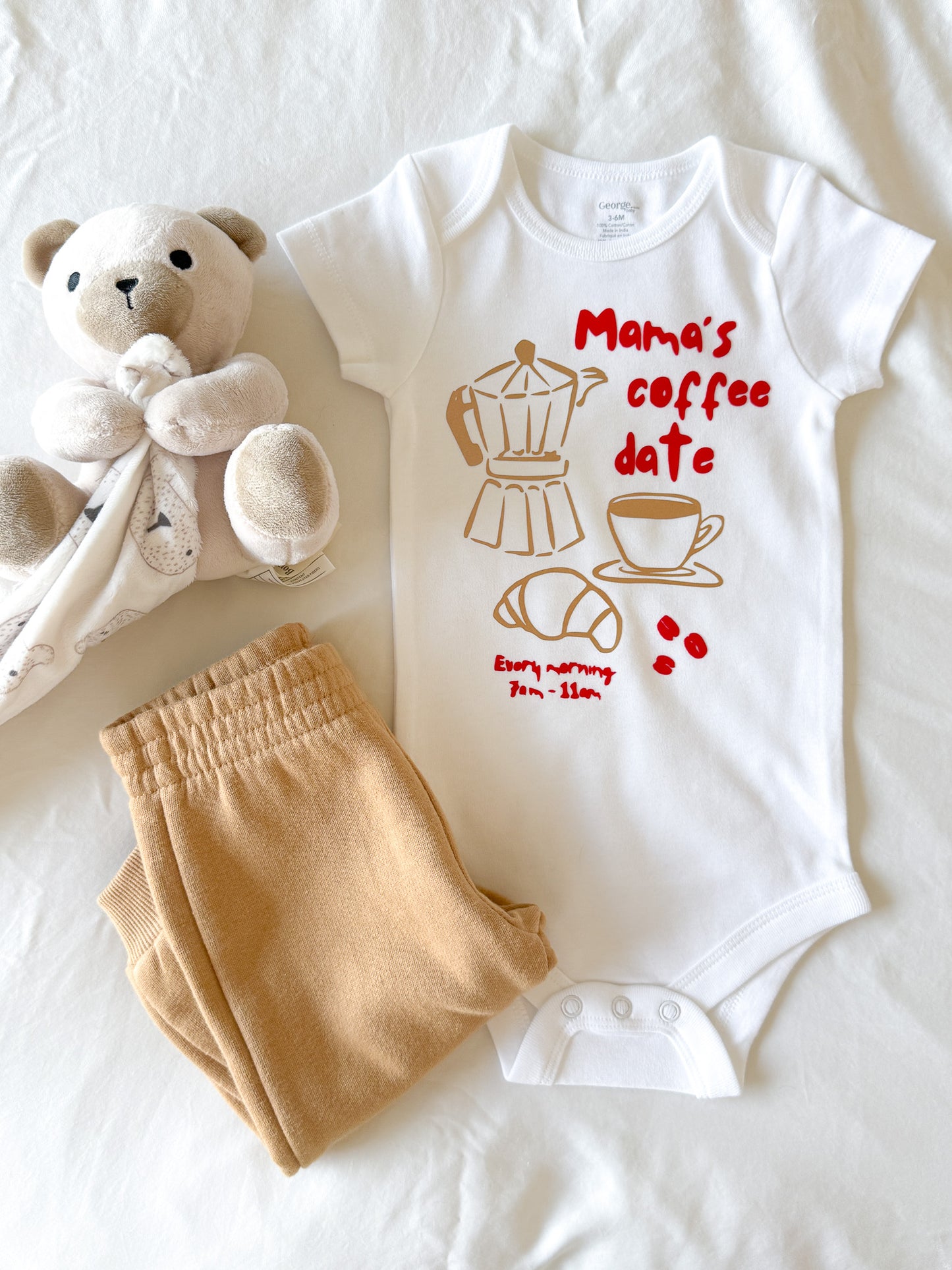 Baby onesie with 'Mama's coffee date' print, beige pants, and teddy bear on a white background