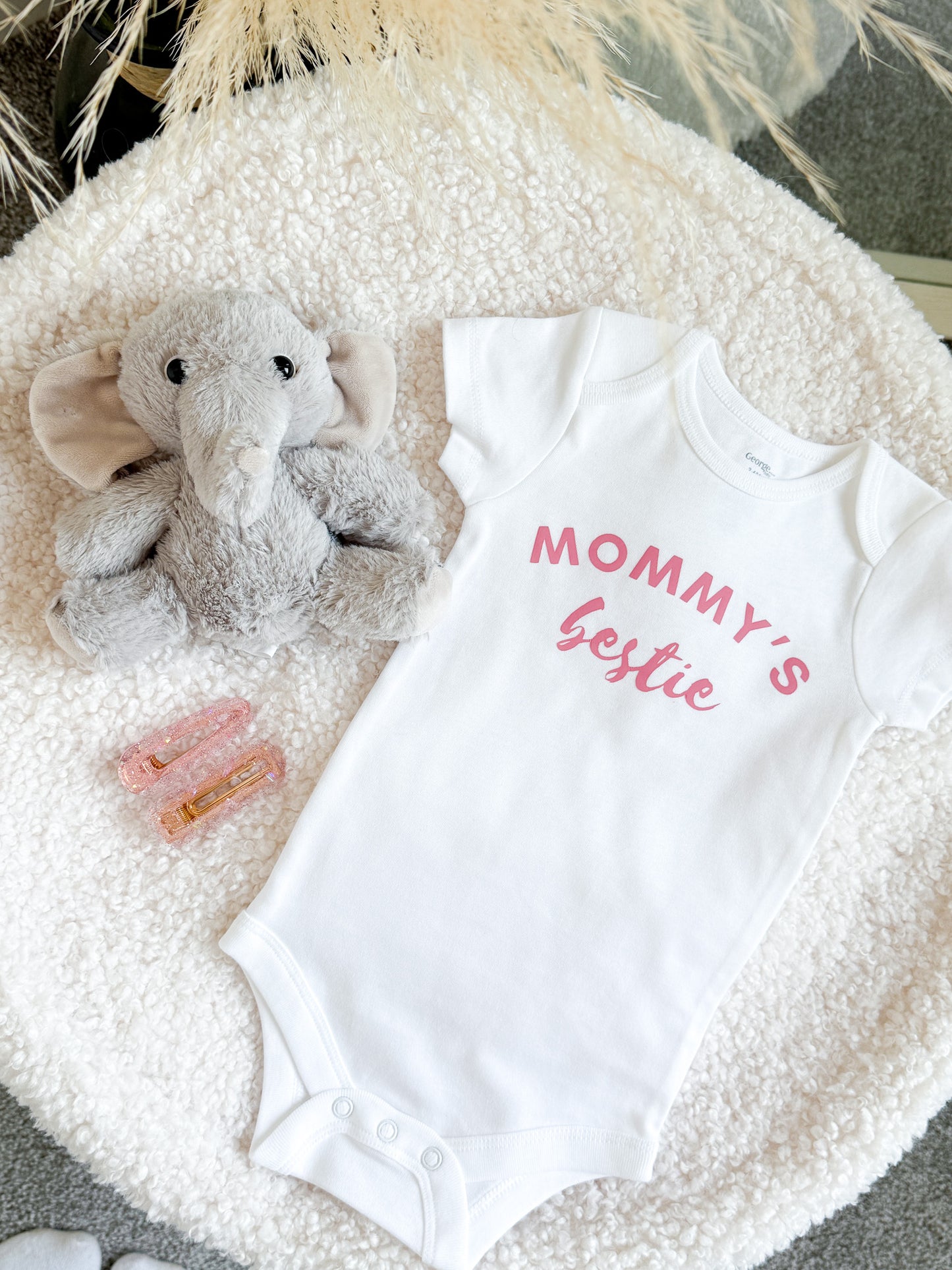 White baby onesie with 'Mommy's Bestie' text, plush elephant toy, and hair clips on a textured surface.