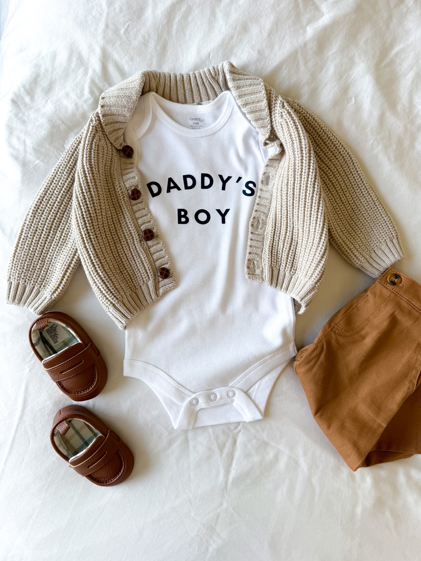 Baby outfit with white 'DADDY'S BOY' onesie, beige cardigan, brown pants, and shoes on a white background