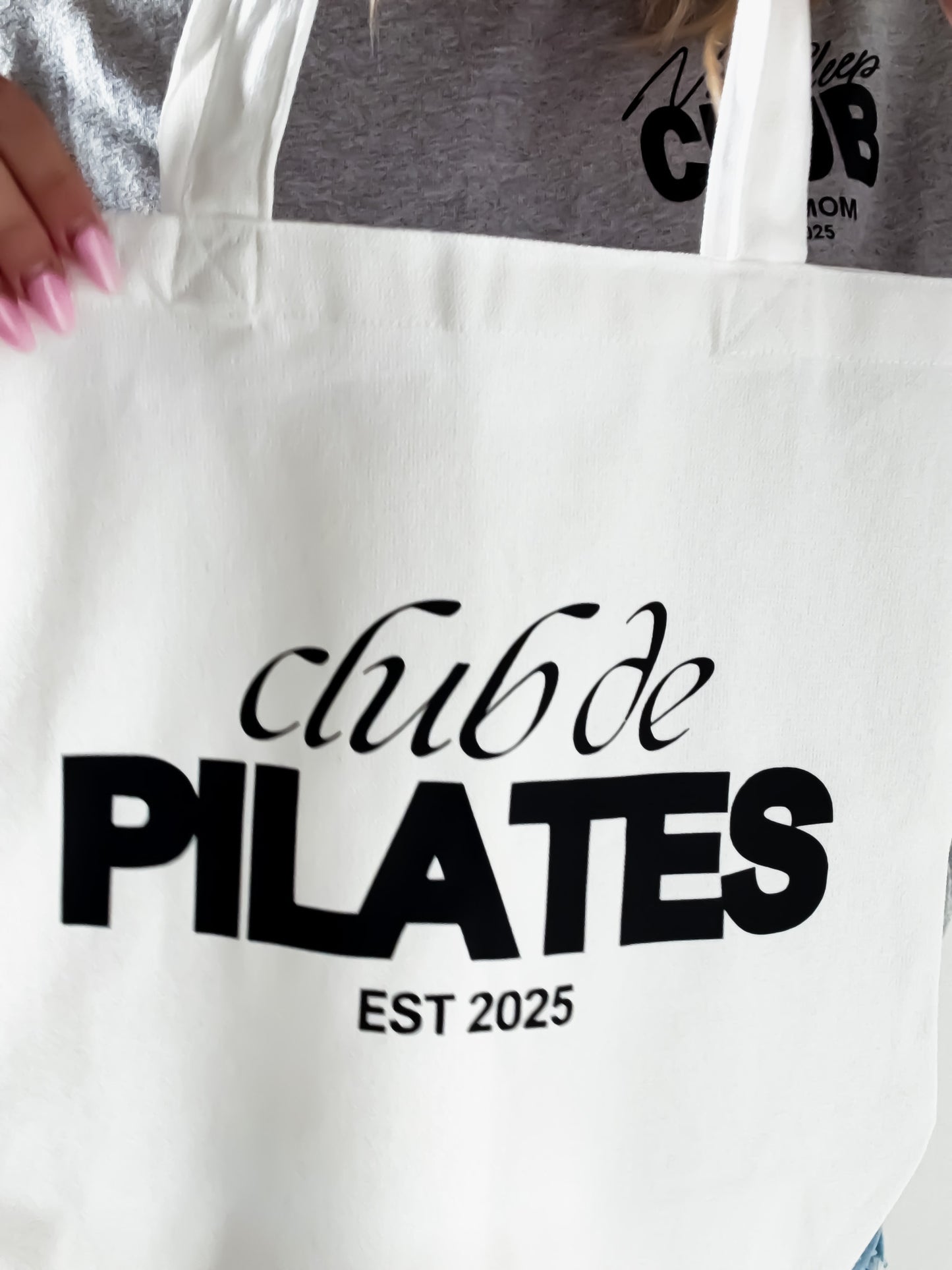 White tote bag with 'club de PILATES EST 2025' text held by a person wearing a gray shirt with similar text.