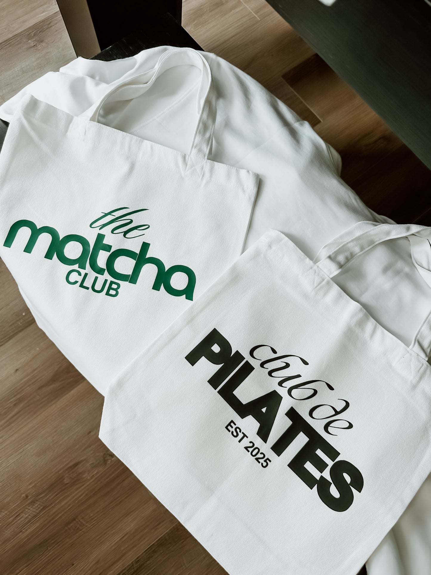 Two white tote bags with 'The Matcha Club' and 'Pilates' text on a wooden surface.