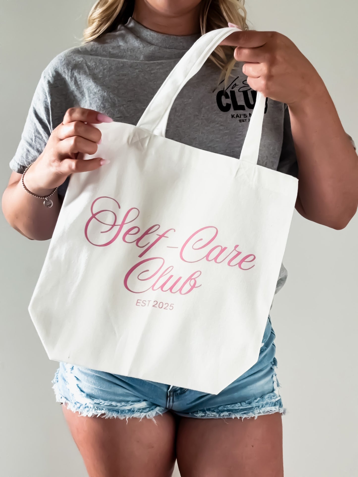 Person holding a white tote bag with 'Self Care Club' text.