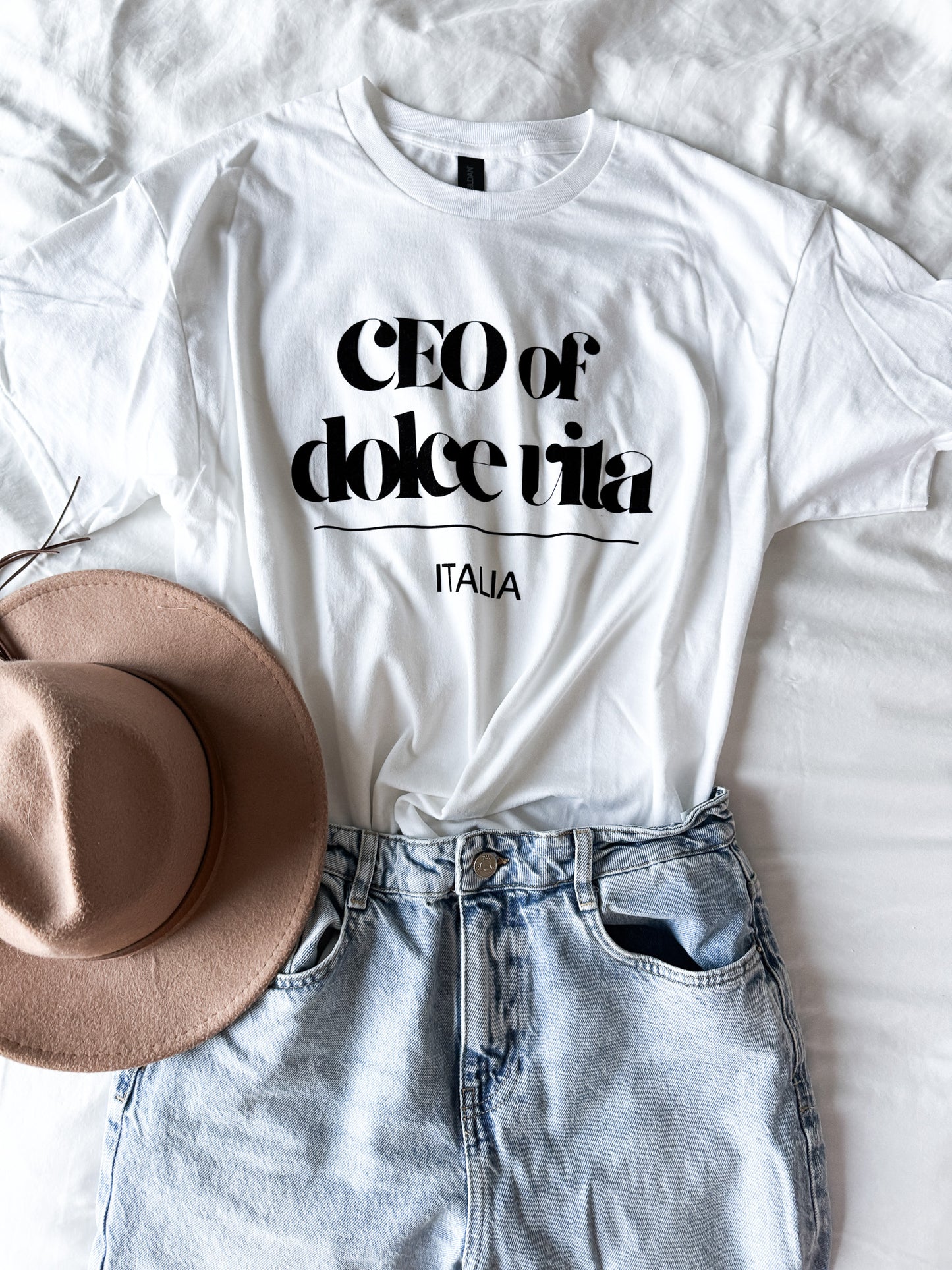 White t-shirt with 'CEO of dolce vita Italia' text, paired with a beige hat and denim shorts on a white background.