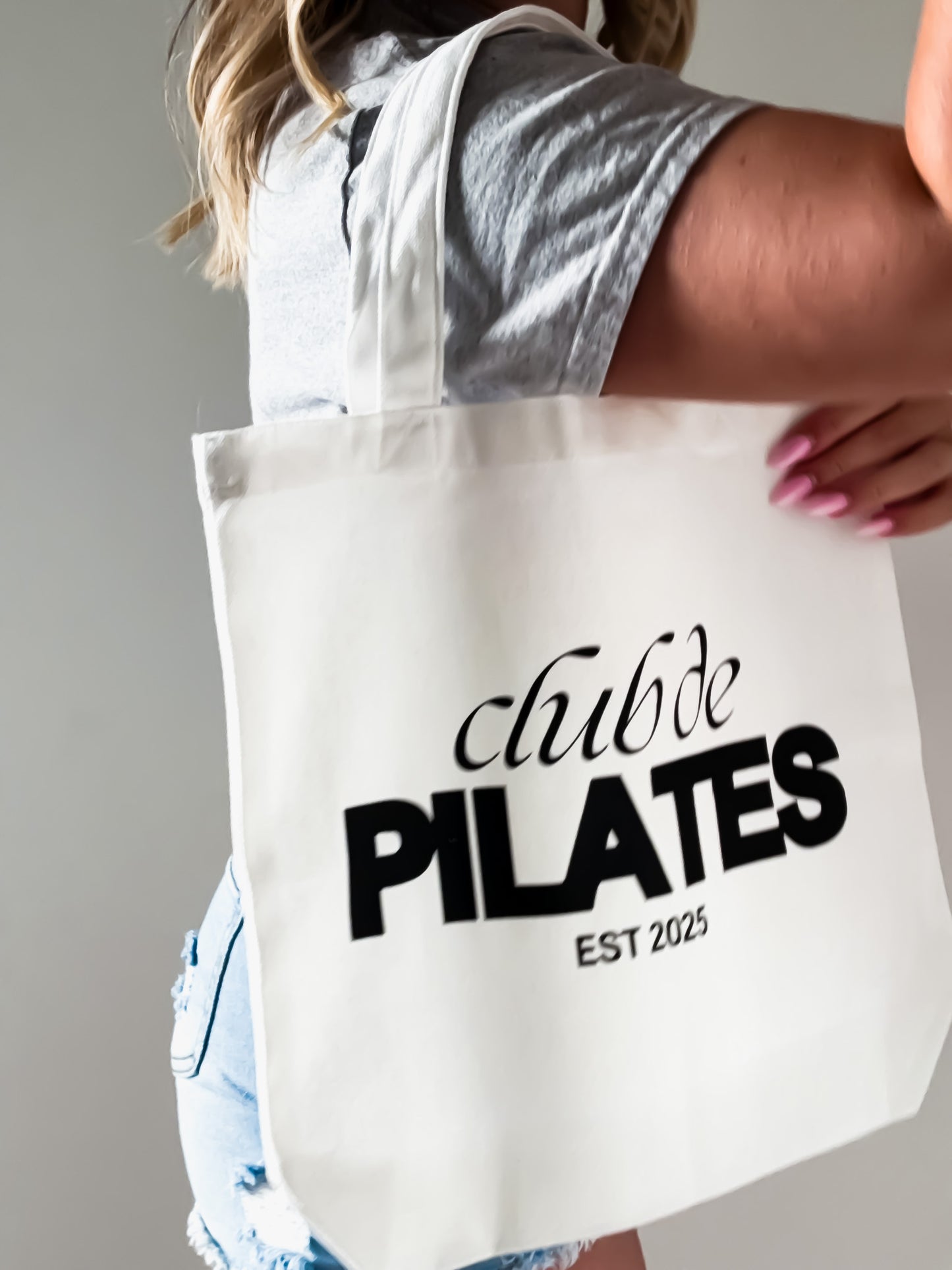 Person holding a tote bag with 'club de PILATES EST 2025' text.