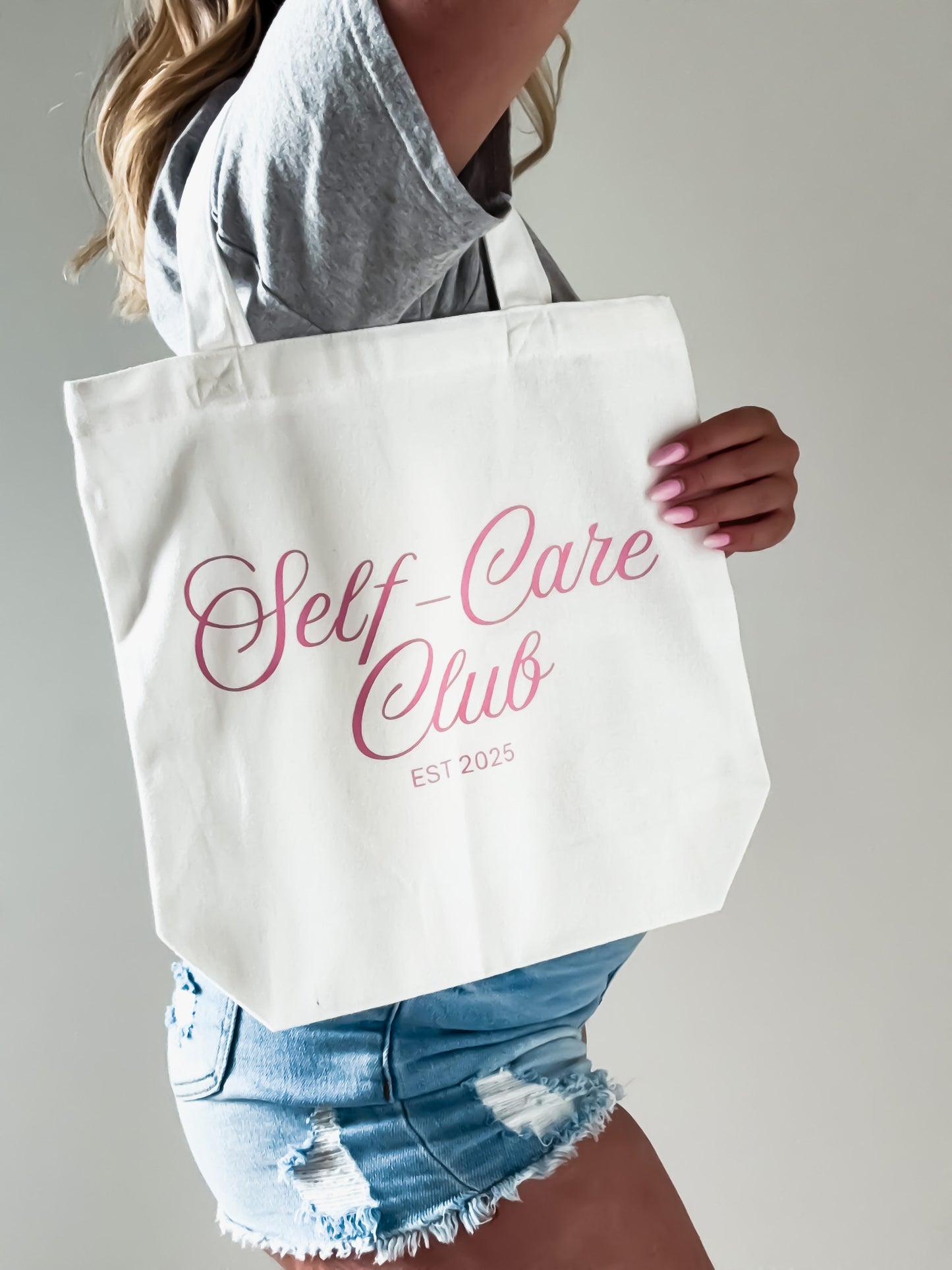 Person holding a tote bag with 'Self-Care Club' text on a plain background