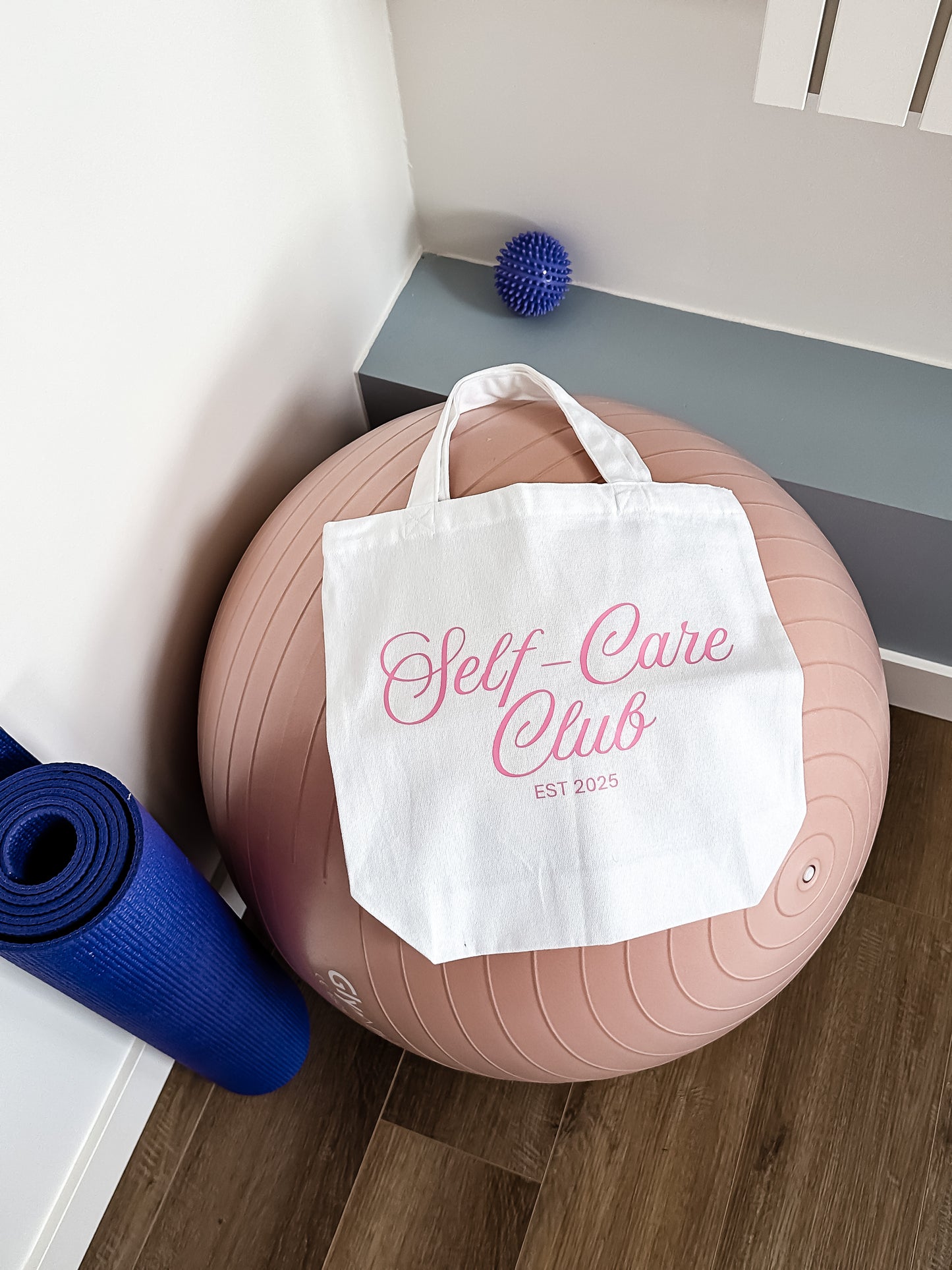 White tote bag with 'Self-Care Club' text on a pink exercise ball next to a blue yoga mat.
