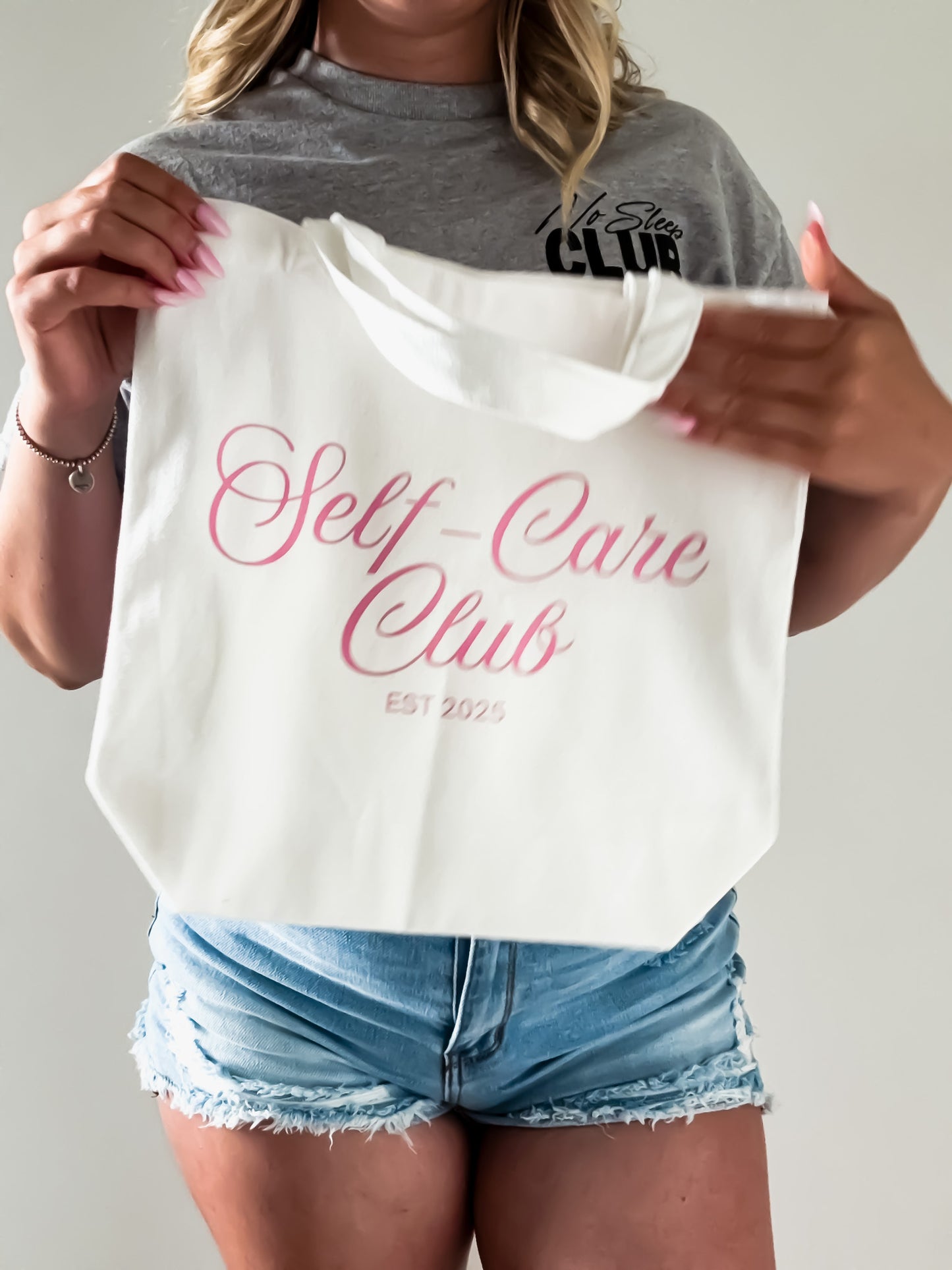 Person holding a white tote bag with 'Self-Care Club' text, wearing a gray t-shirt and denim shorts.