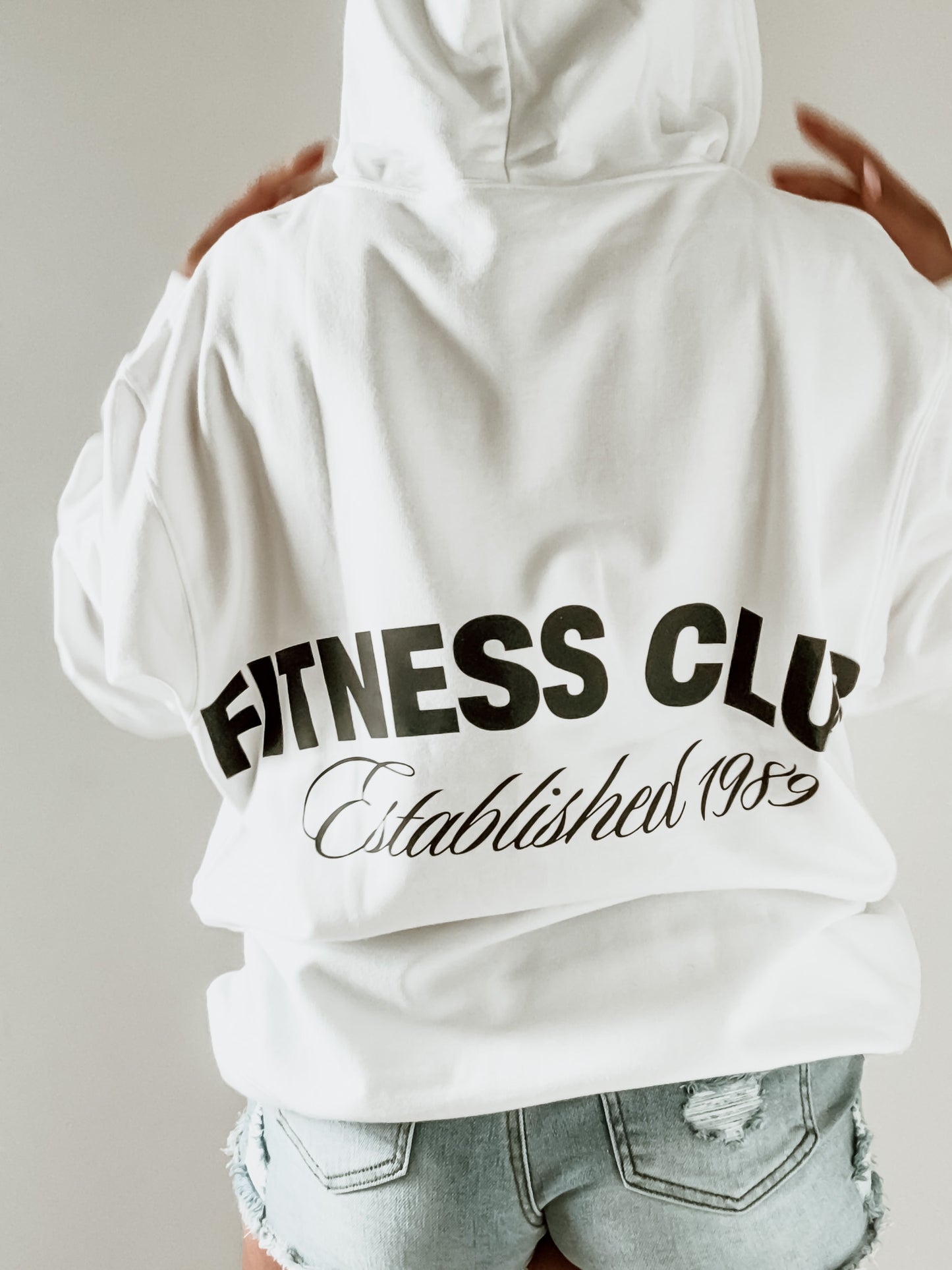 White hoodie with 'Fitness Club Established 1985' text on the back, worn by a person.