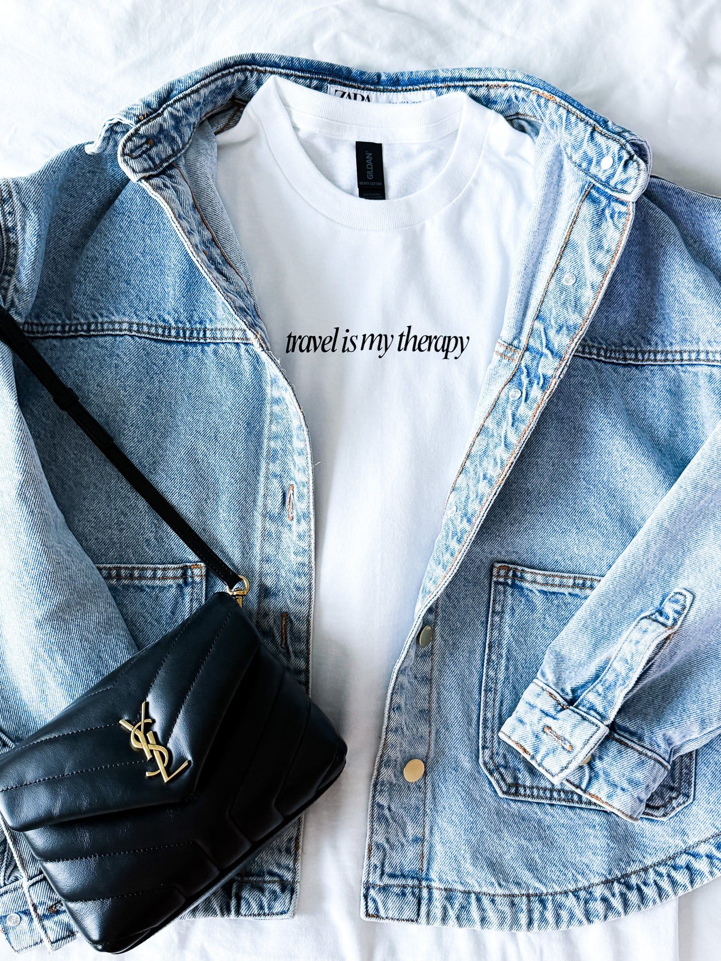 White t-shirt with 'travel is my therapy' text, denim jacket, and black handbag on a white background