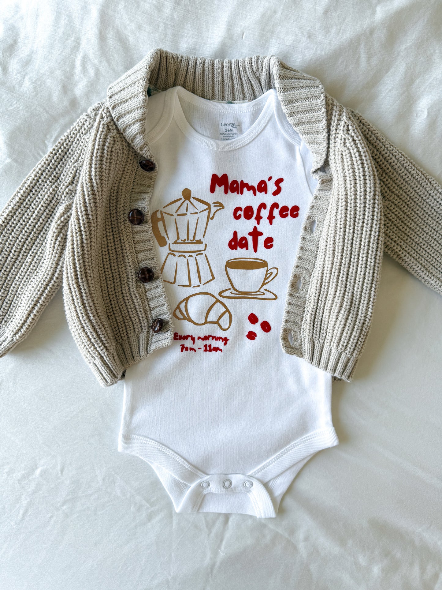 Baby onesie with 'Mama's coffee date' text and graphics, paired with a knitted cardigan on a white background.