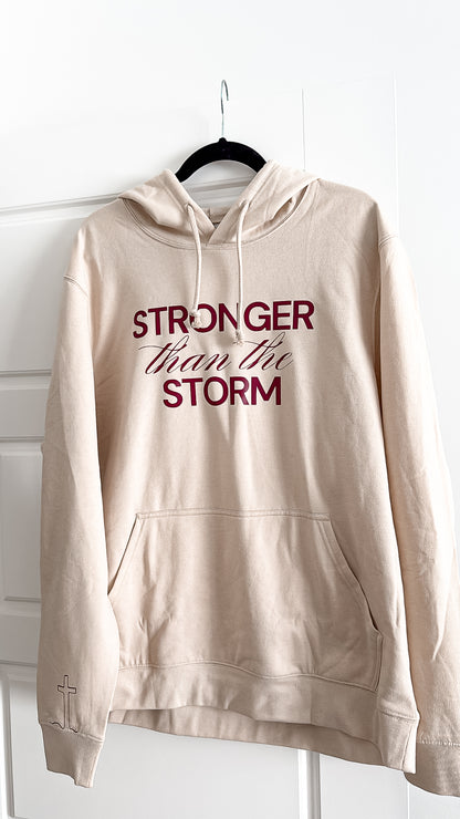 Beige hoodie with 'Stronger than the Storm' text on a hanger against a white background