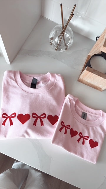 Two pink children's shirts with red bows and hearts on a white surface.