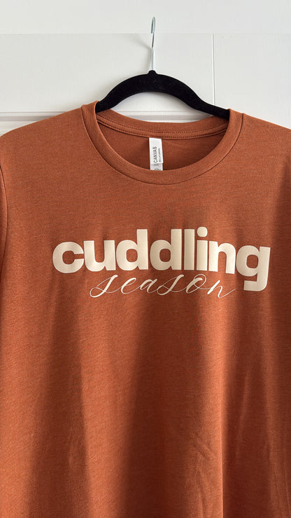 Brown t-shirt with 'cuddling season' text on a hanger against a white wall.