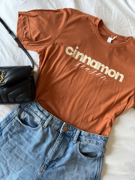 burnt orange t-shirt with 'cinnamon season' text, blue jeans, and black handbag on a bed