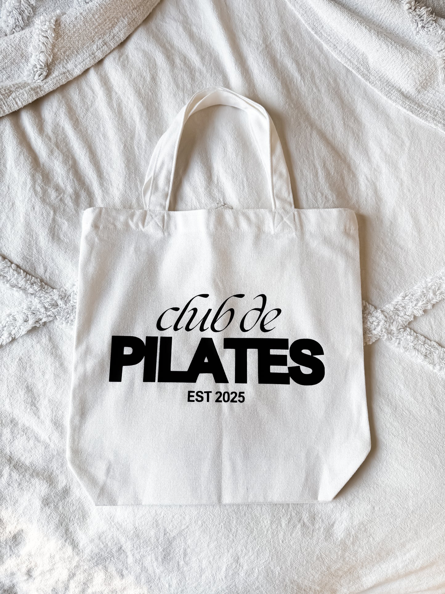 White tote bag with 'club de PILATES EST 2025' text on a textured surface