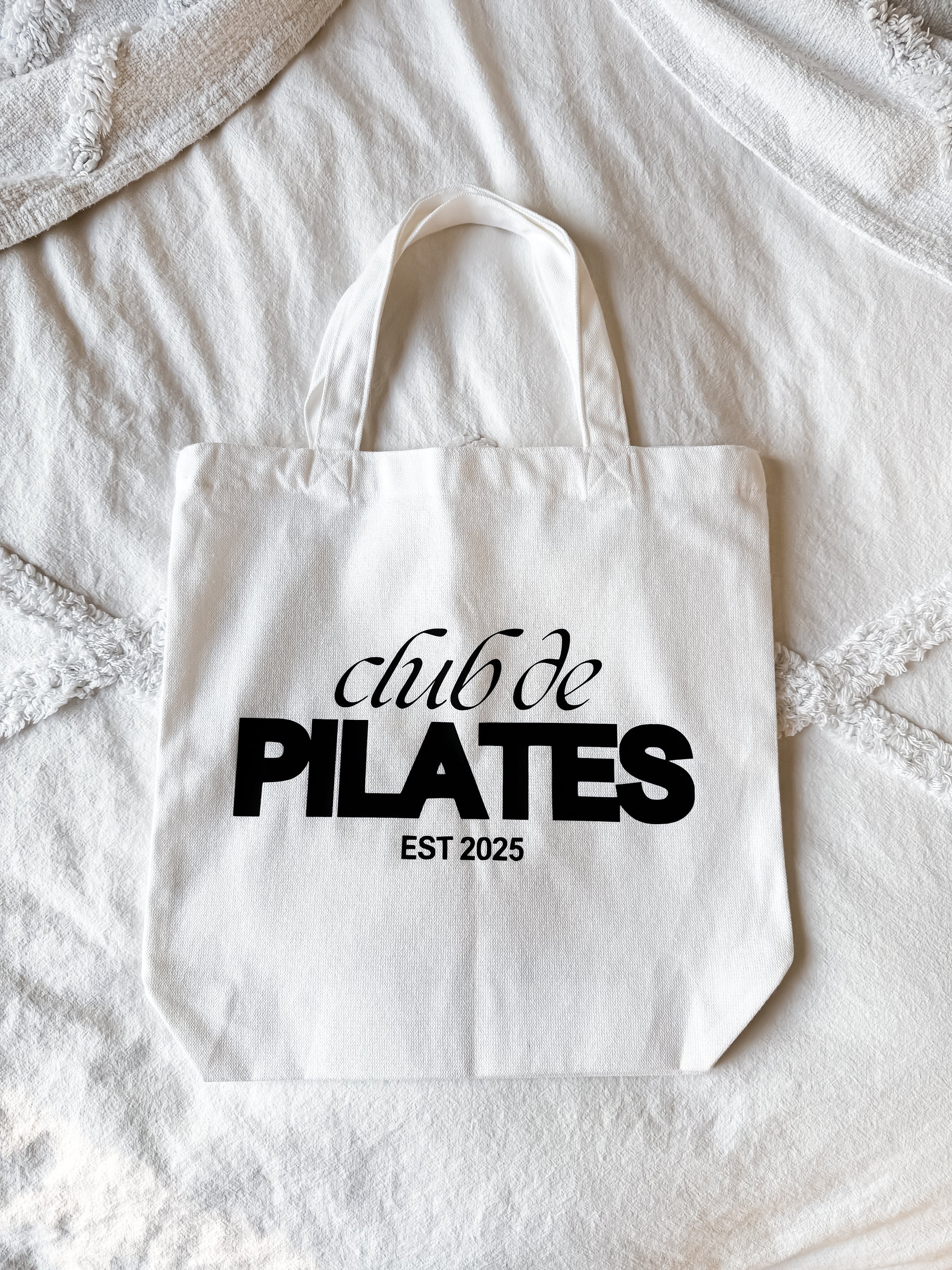 White tote bag with 'club de PILATES EST 2025' text on a textured surface