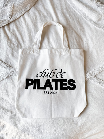 White tote bag with 'club de PILATES EST 2025' text on a textured surface