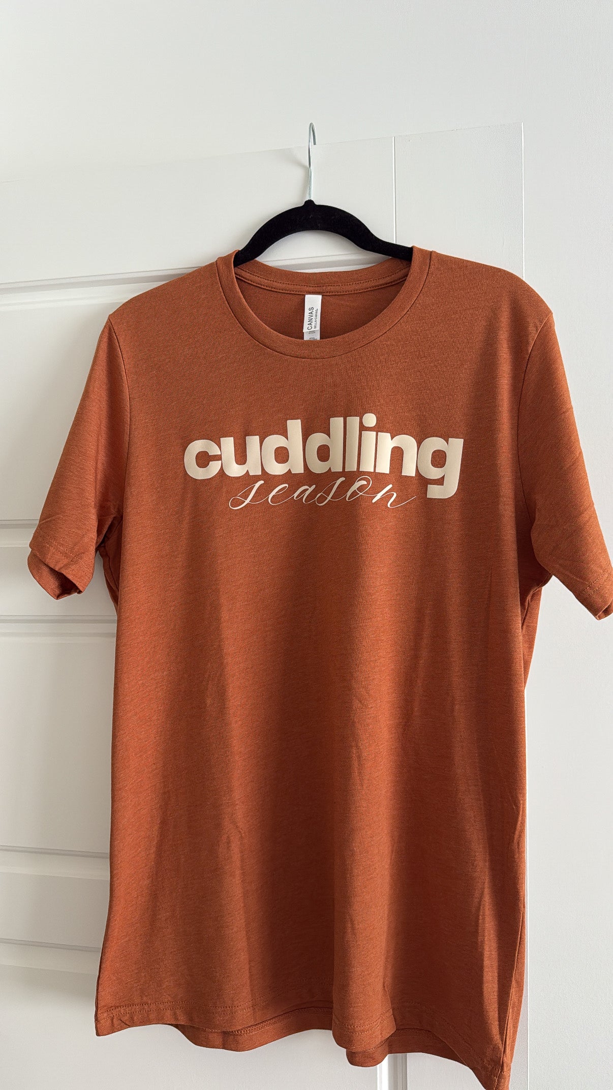 Brown t-shirt with 'cuddling season' text on a hanger against a white wall.