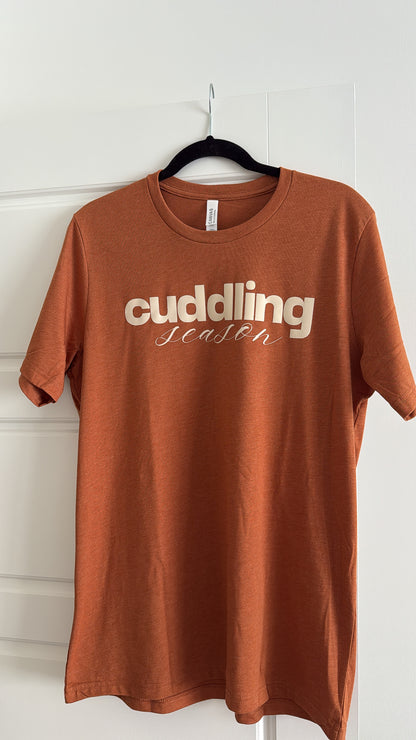 Brown t-shirt with 'cuddling season' text on a hanger against a white wall.