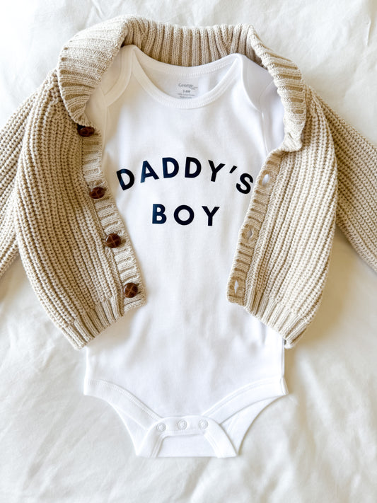 White baby onesie with 'DADDY'S BOY' text and beige cardigan on a white background