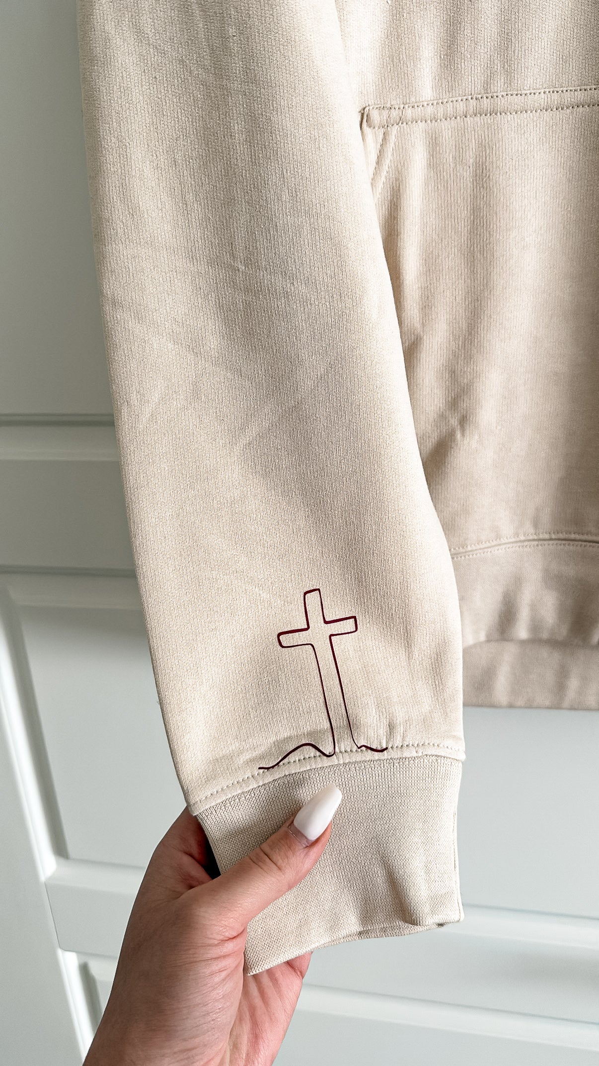 Beige sweatshirt with a cross design held by a hand against a white background