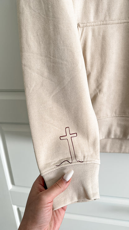 Beige sweatshirt with a cross design held by a hand against a white background