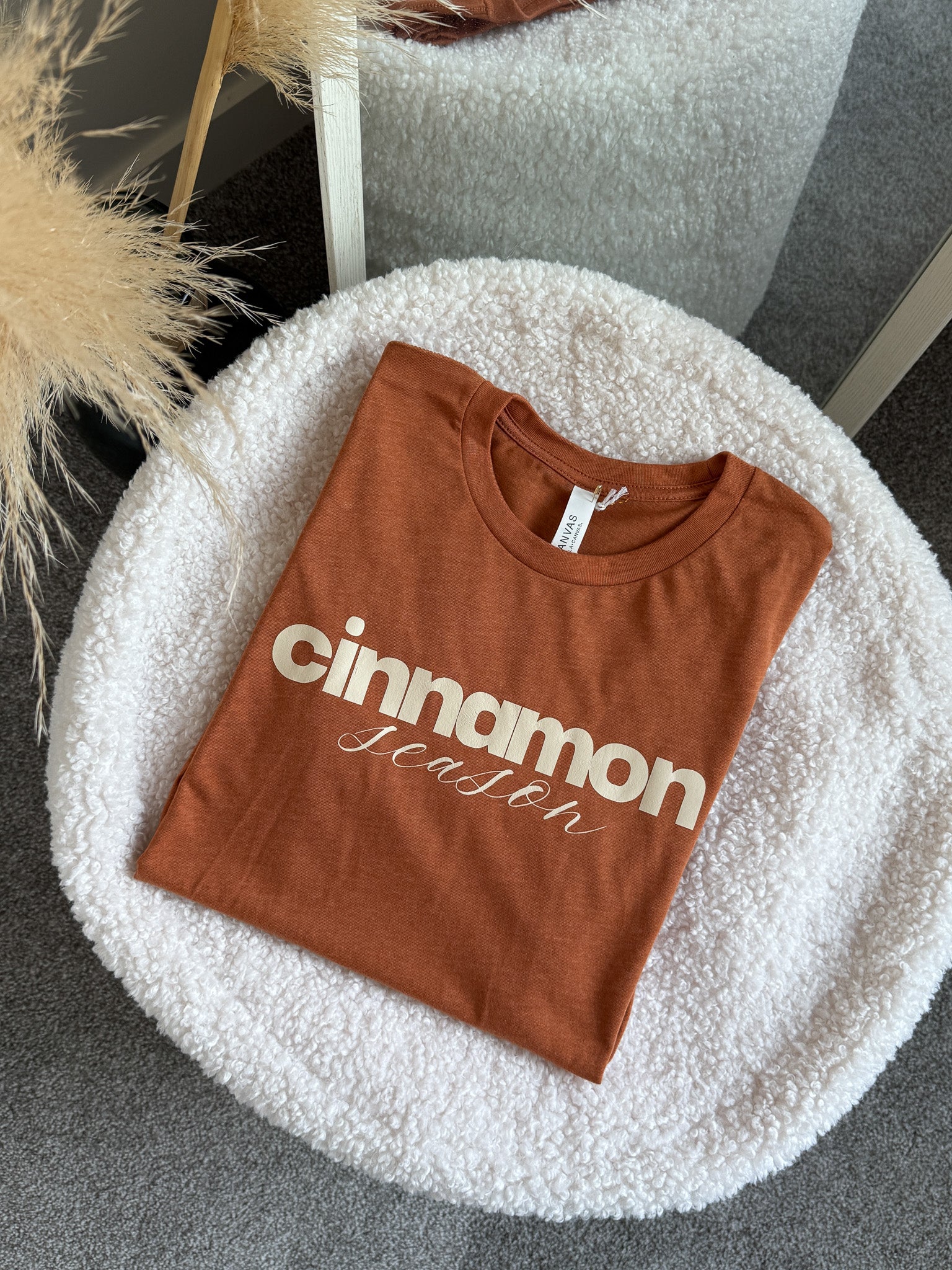 Folded brown t-shirt with 'cinnamon season' text on a white round surface.