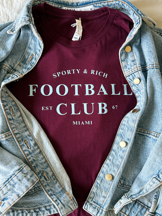 Maroon t-shirt with 'Sporty & Rich Football Club' text, worn over a denim jacket.