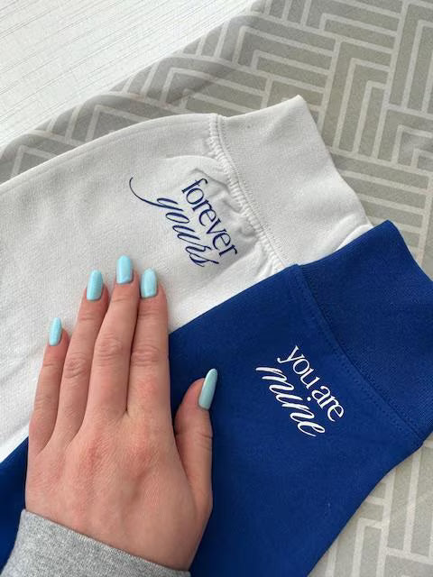 Two sweatshirts, one white with blue text and one blue with white text, held together by a hand.