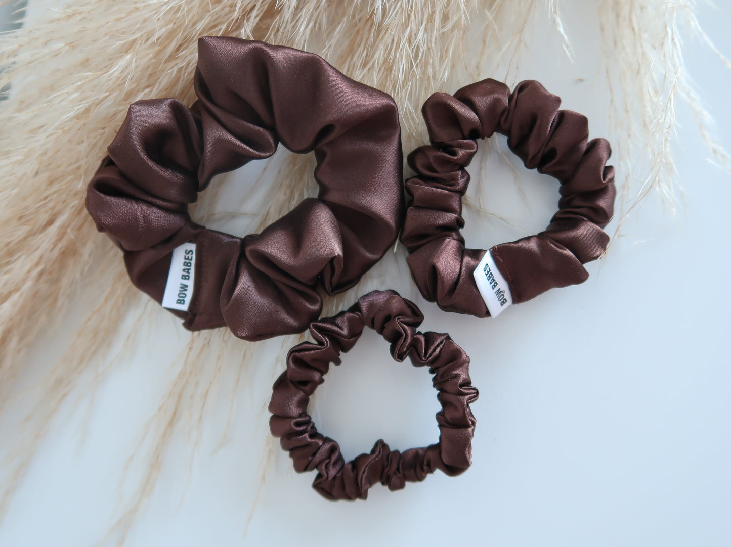 set of three chocolate brown scrunchies on white backdrop with wheat grass in background