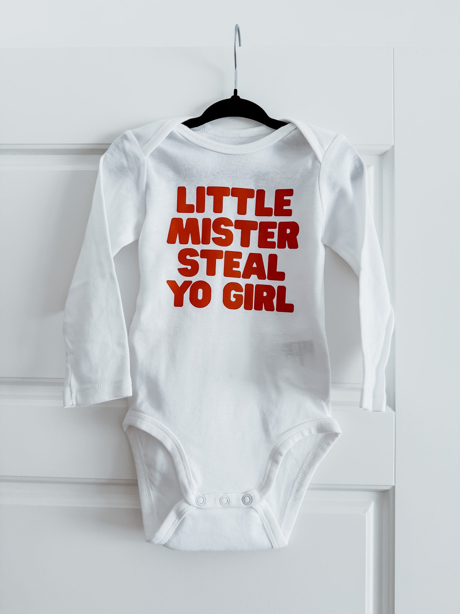 White baby onesie with red text on a hanger against a white wall