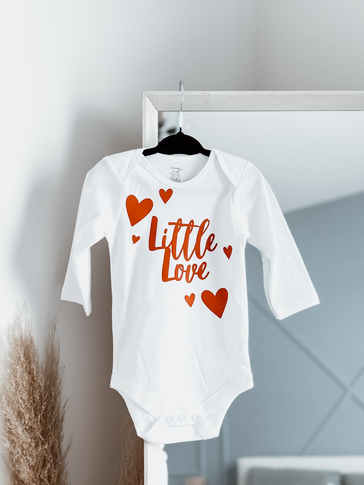 White baby onesie with red text and hearts on a hanger against a light gray wall.