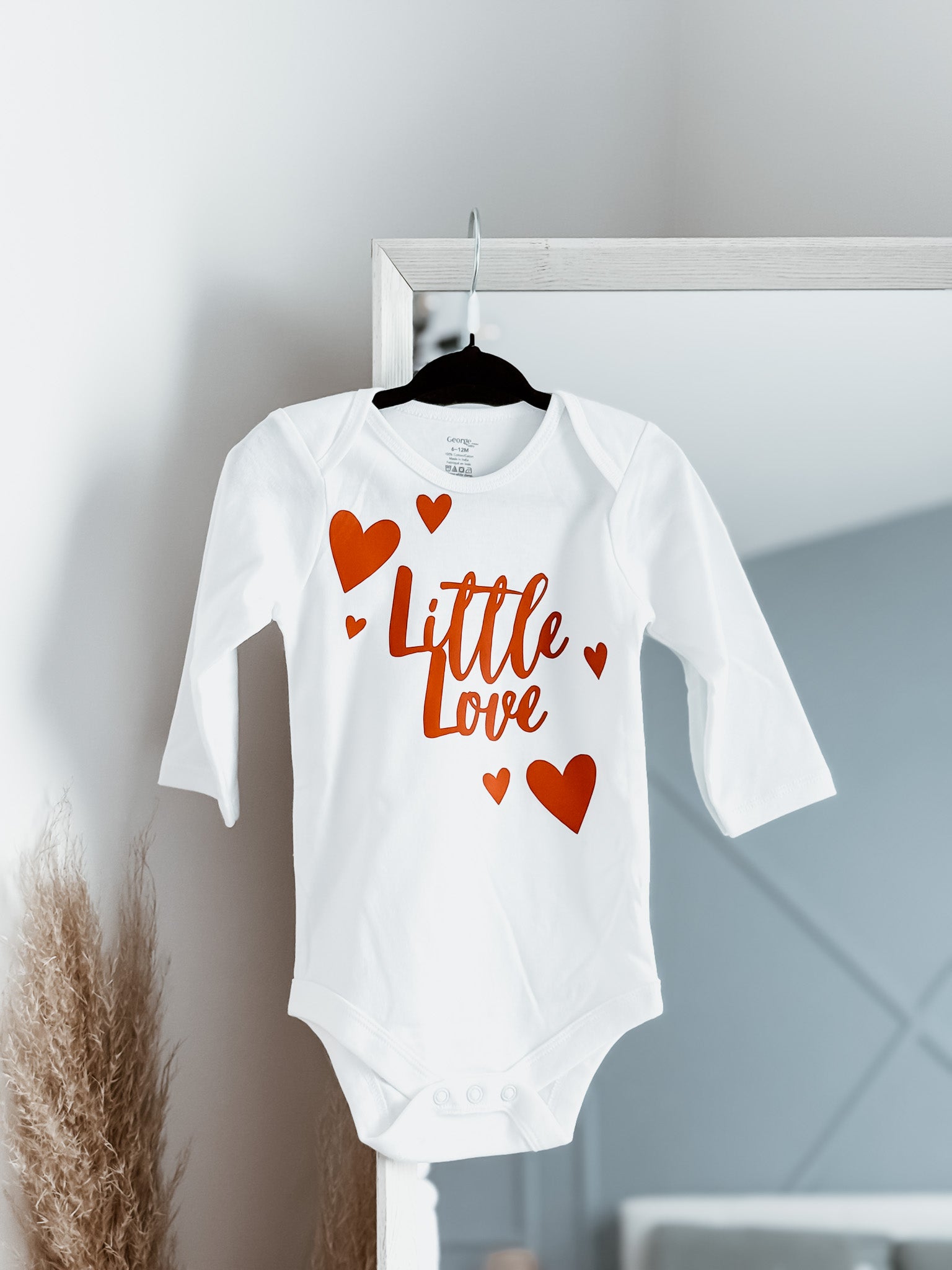 White baby onesie with red text and hearts on a hanger against a light gray wall.