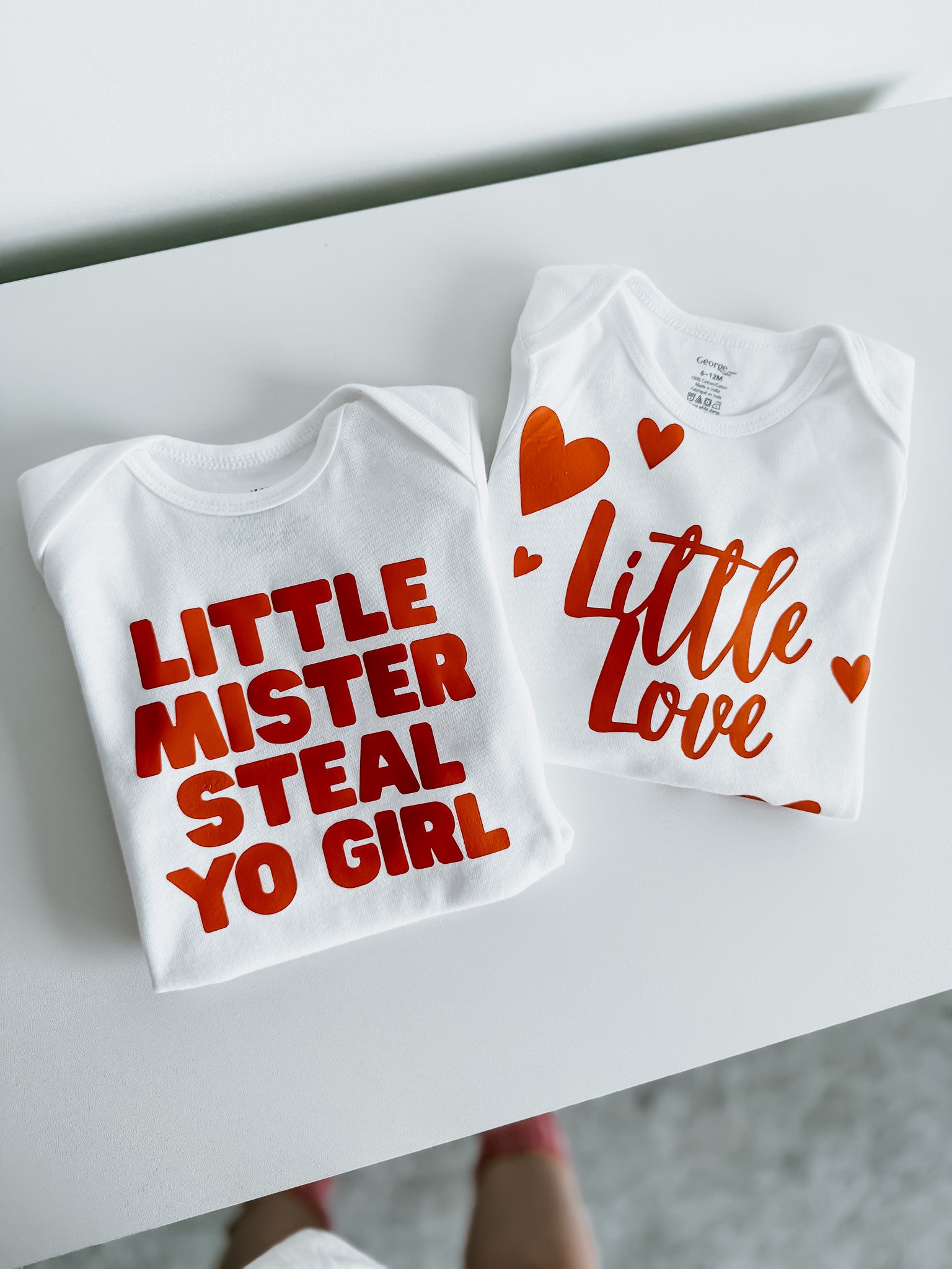 Two white baby onesies with red text on a white surface