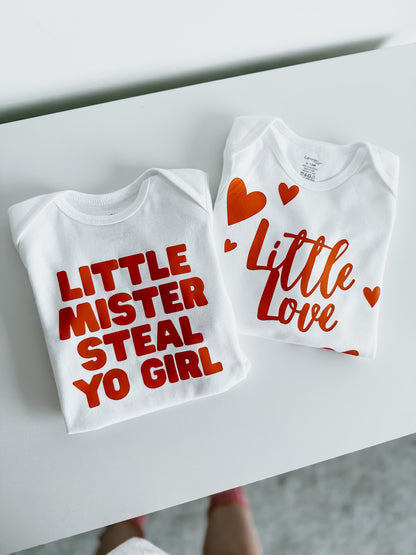 Two white baby onesies with red text on a white surface