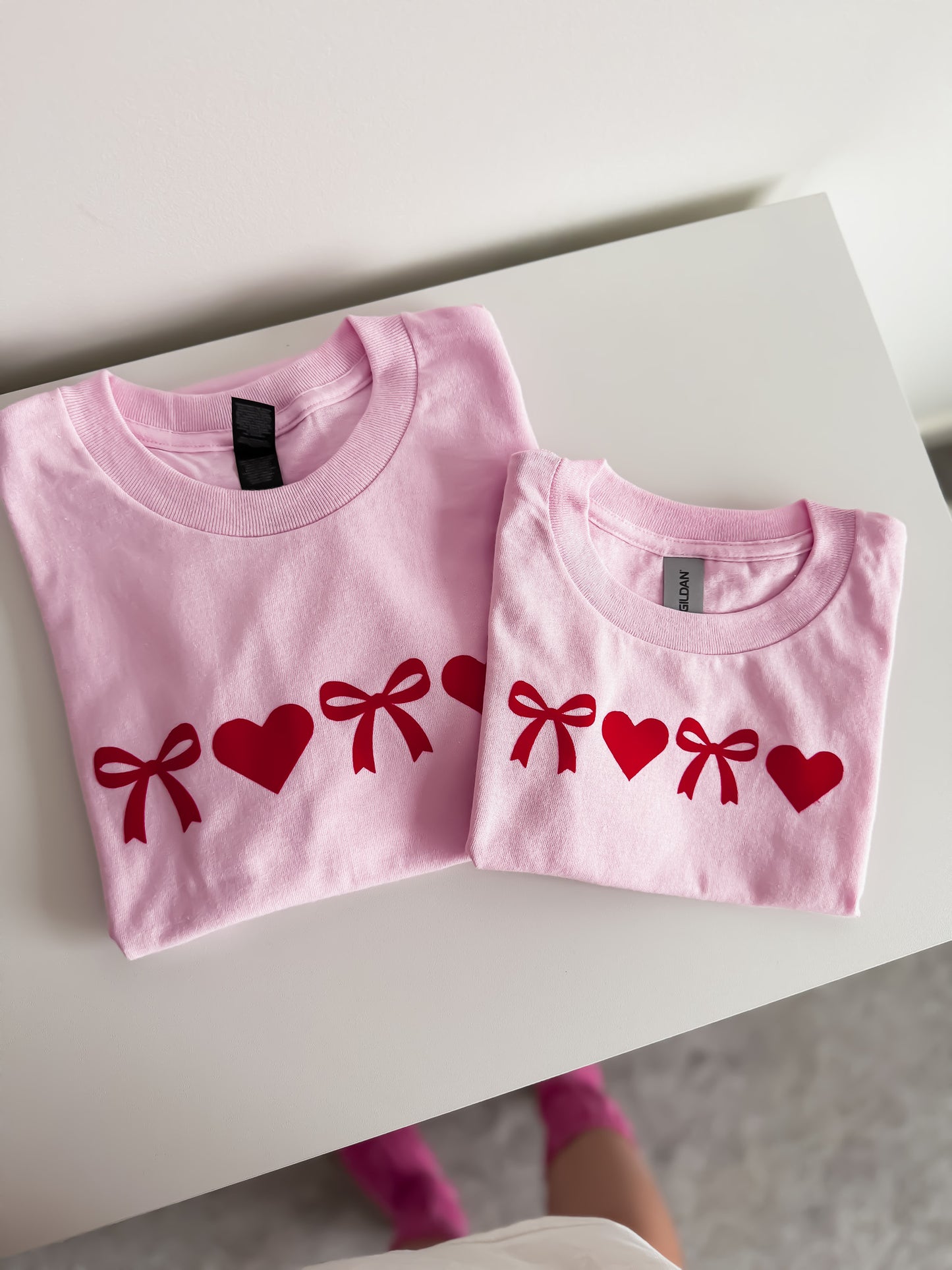 Two pink t-shirts with red heart and bow designs on a white surface.