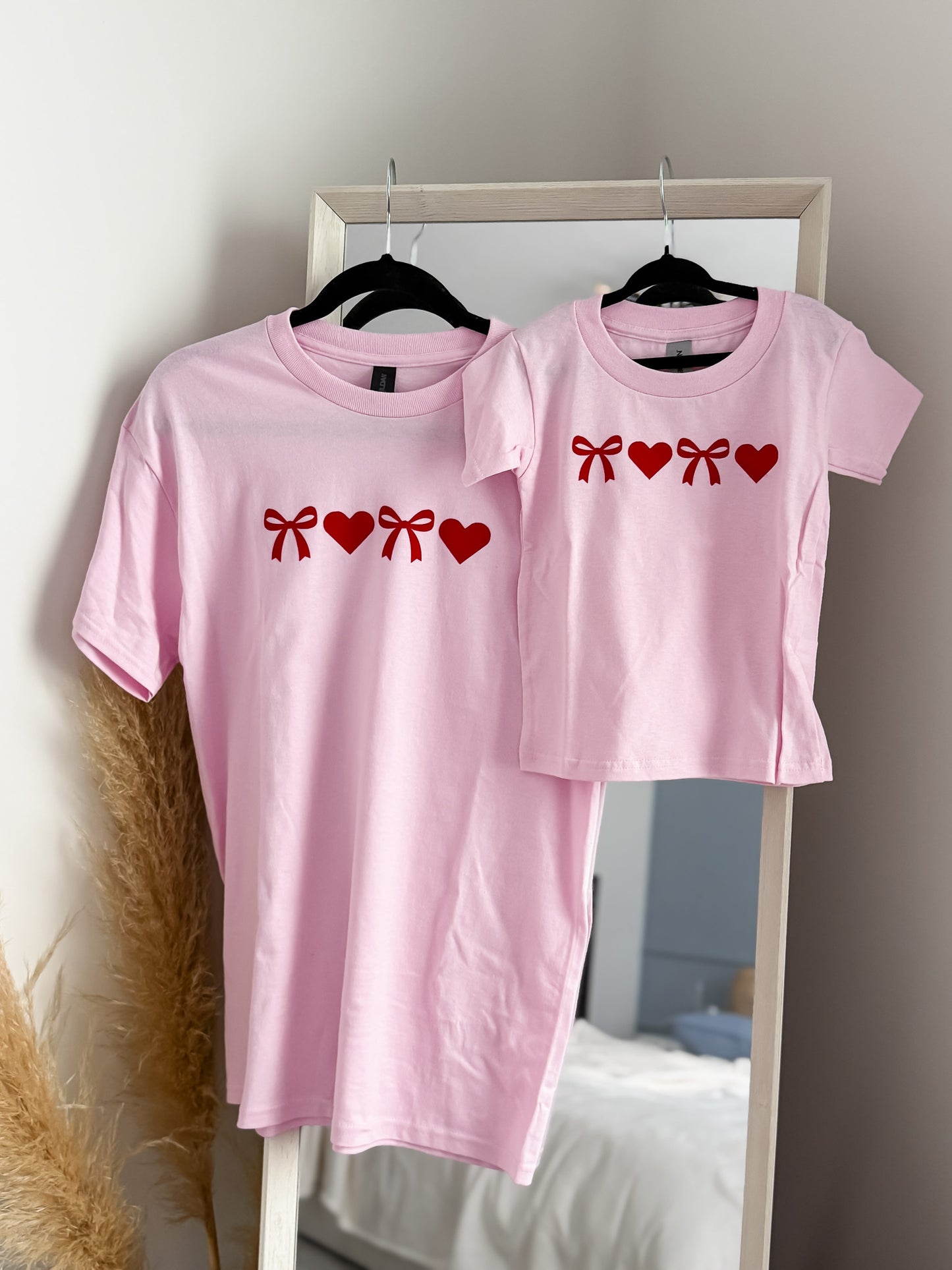 Two pink t-shirts with red graphics on a hanger against a neutral wall.