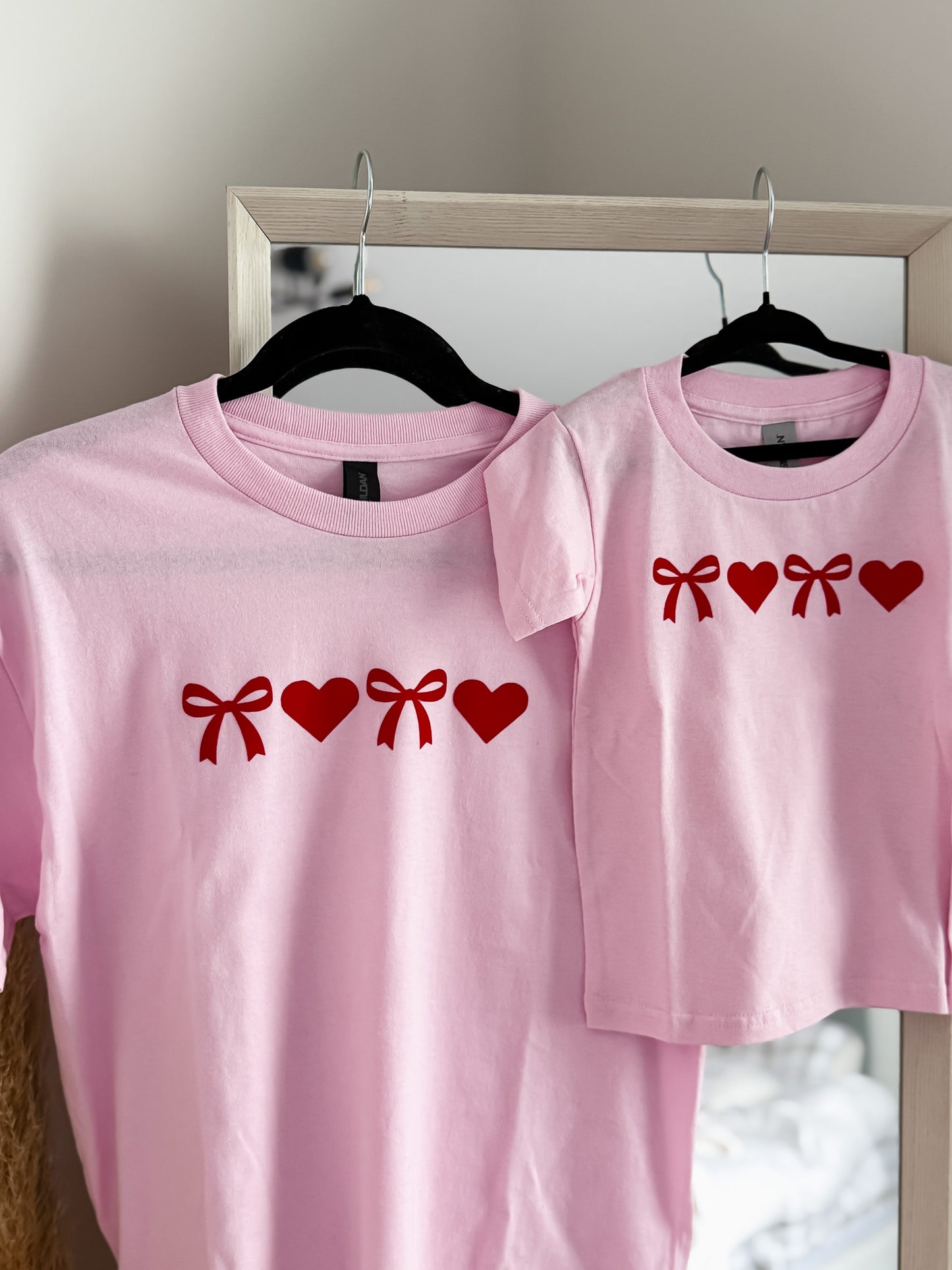 Two pink t-shirts with red bow and heart designs on hangers against a neutral background.