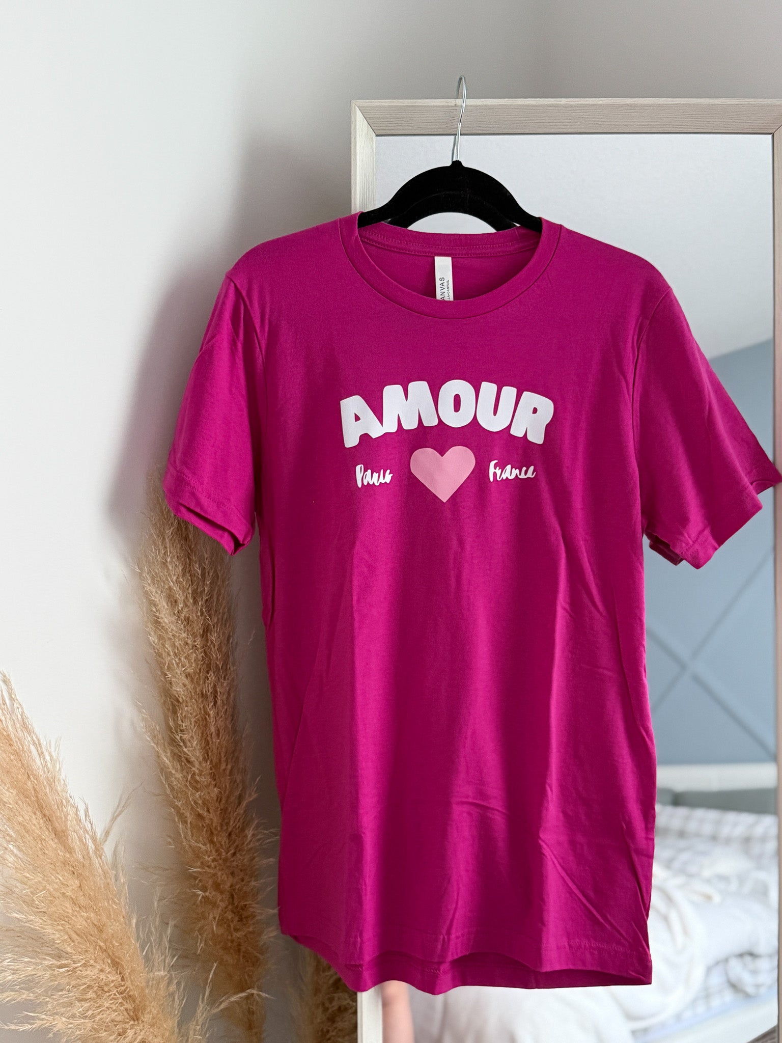 Pink t-shirt with 'AMOUR' text and heart graphic on a hanger against a neutral wall.