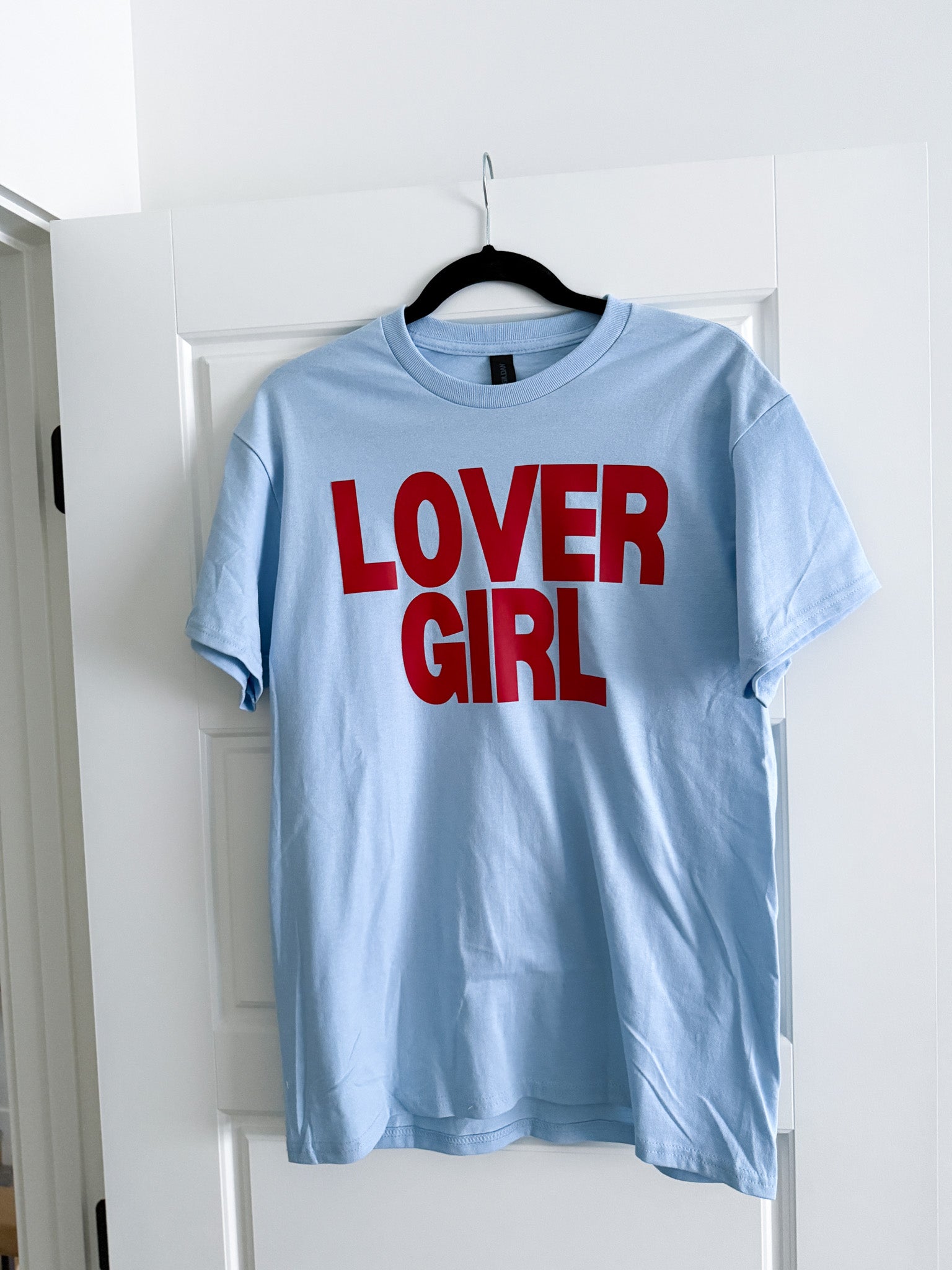 Light blue t-shirt with 'LOVER GIRL' in red text on a hanger against a white door.