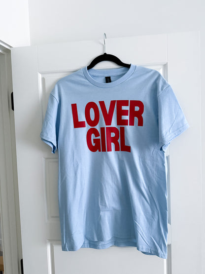 Light blue t-shirt with 'LOVER GIRL' in red text on a hanger against a white door.